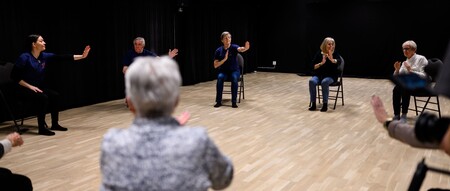 En grupp äldre personer sitter i en cirkel på stolar och gör rörelseövningar i ett ljust rum med trägolv.