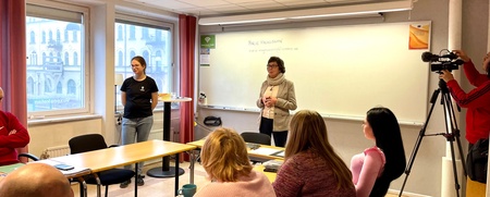 En grupp människor sitter i ett klassrum medan en kvinna står och pratar framför en whiteboard.