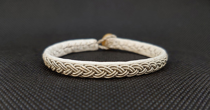 Ett armband med flätad metalltråd och ljust band ligger på en mörk texturerad yta.