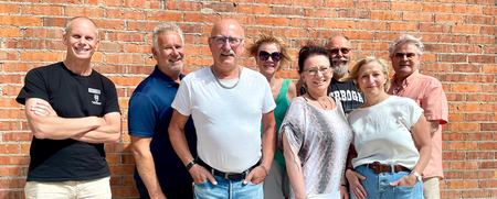 Åtta vuxna personer står tillsammans framför en röd tegelvägg utomhus, leende mot kameran.