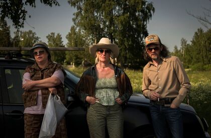 Tre personer står framför en bil på landet, klädda i västerninspirerade kläder.