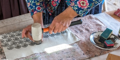 En person trycker mönster på tyg med hjälp av en stämpel och hammare vid ett bord.