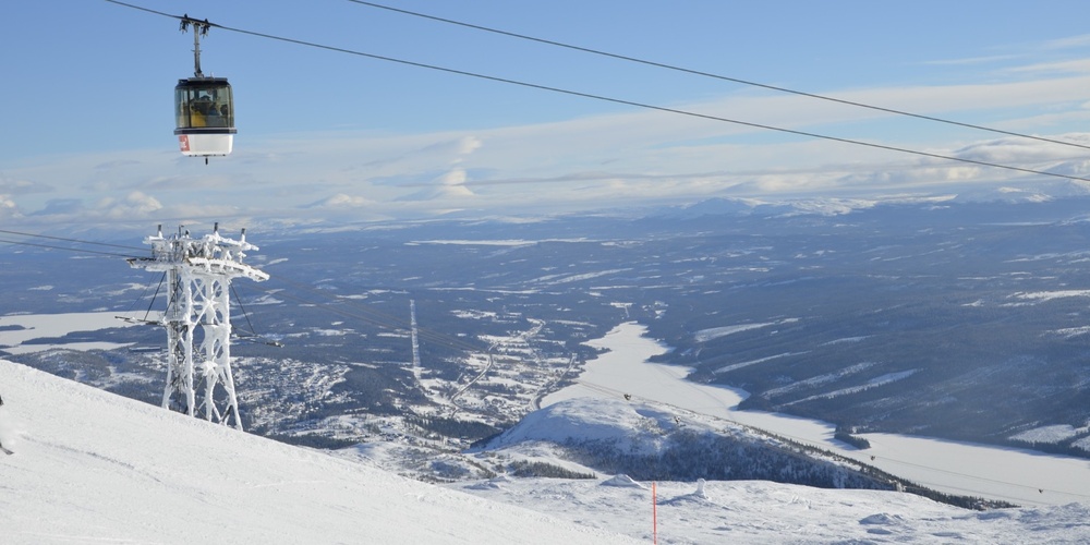 Landskapsvy över snötäckta fjäll med en linbana och liftstolpe, samt sjöar och skog i bakgrunden.