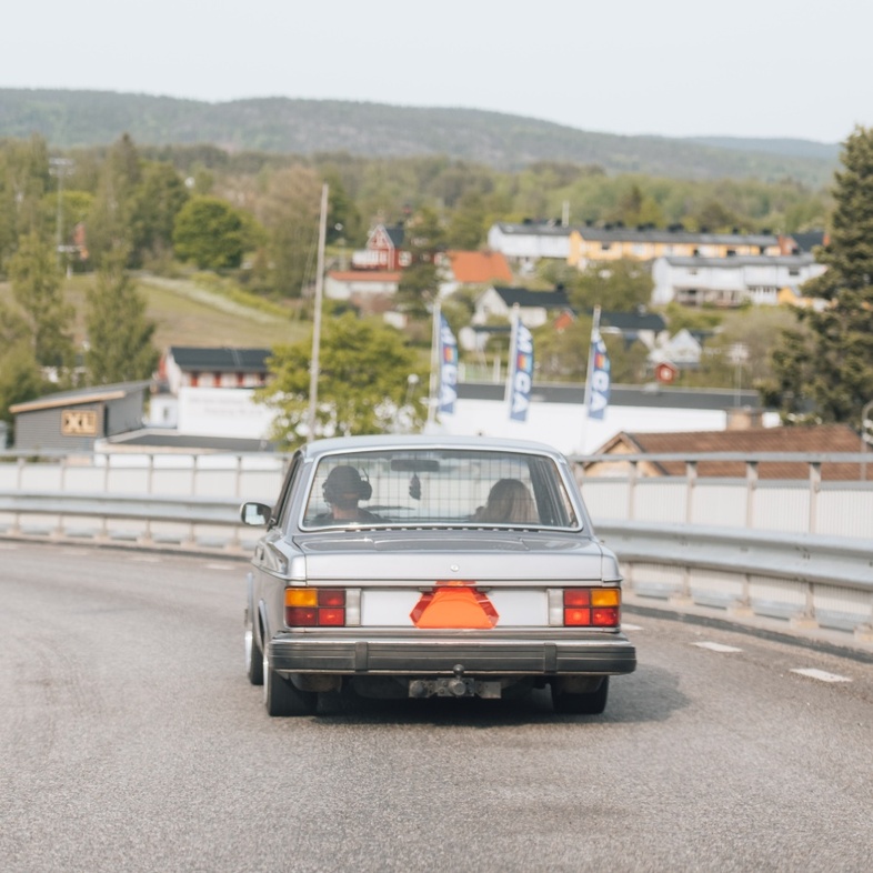 En äldre silverfärgad bil ombyggd till Epa-traktor kör på en landsväg med två personer inuti, landsbygd och hus i bakgrunden.