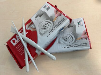 Två paket lufttorkande lera och tre vita modelleringsverktyg på ett ljust bord.