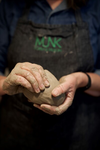 En person formar lera med händerna, iförd förkläde med texten 'MUNK'.