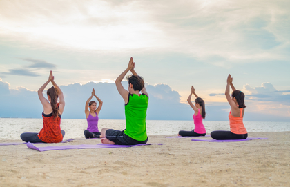 Fem personer gör yoga på stranden vid havet i solnedgången, sittande i en cirkel med händerna ovanför huvudet.