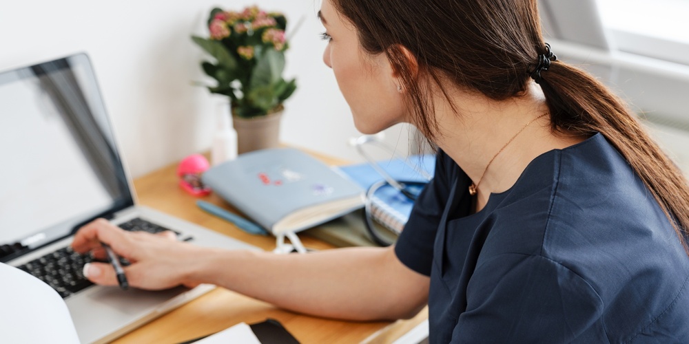 Kvinna i blå arbetskläder arbetar vid skrivbord med laptop och papper, sedd bakifrån.