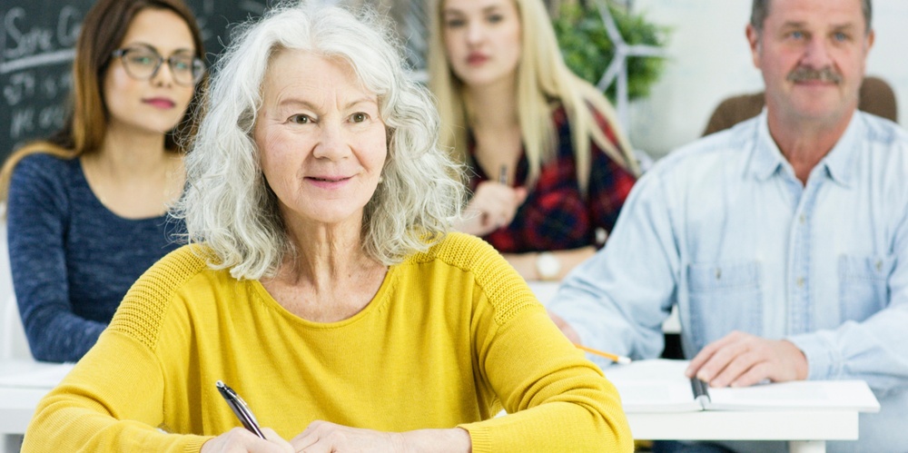 Äldre kvinna i gul tröja sitter i klassrum med andra vuxna och antecknar.
