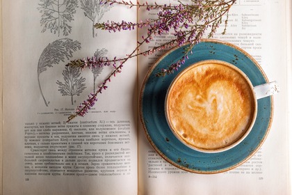 En kopp cappuccino på ett blått fat står på en uppslagen bok med botaniska illustrationer och torkade blommor.
