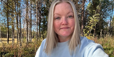 Kvinna med ljust hår och ljus tröja sitter utomhus i en solig skogsmiljö.