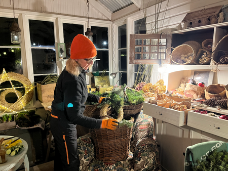 Person med orange mössa och handskar arrangerar grönska i en korg i ett mysigt växthus med naturmaterial.