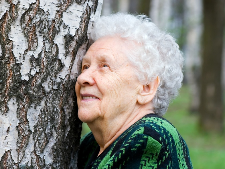 Äldre kvinna med vitt hår lutar sig mot ett träd i en skog och ler.