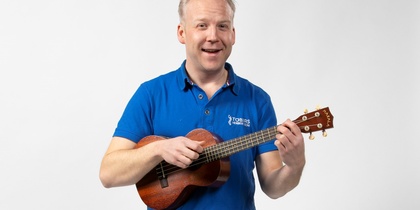 Man i blå pikétröja spelar ukulele och står mot en vit bakgrund.