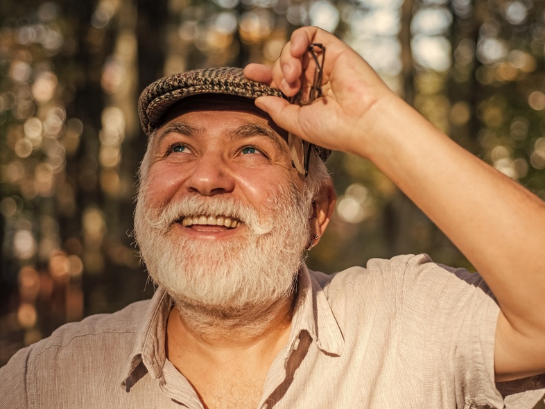 Leende äldre man med vitt skägg och keps står utomhus i soligt skogsparti.