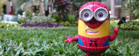 En leksaksfigur som kombinerar en Minion och Spiderman står på gräs i en trädgårdsmiljö.