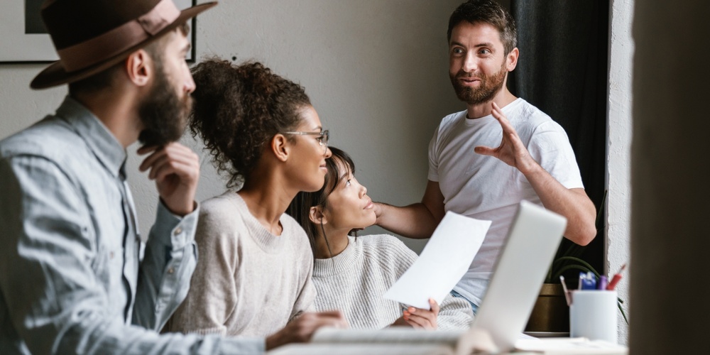 Fyra personer har ett möte vid ett bord, en man står och pratar medan de andra lyssnar.