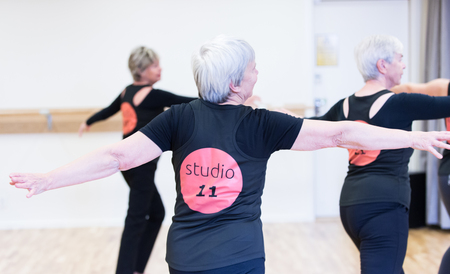 Tre äldre kvinnor dansar i en ljus dansstudio, alla bär svarta kläder med texten 'studio 11' på ryggen.