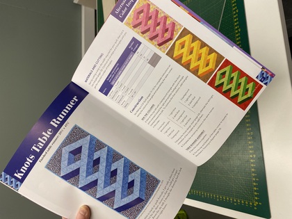 En person håller en öppen bok med instruktioner och bilder på färgglada quiltade bordsdukar i geometriska mönster.