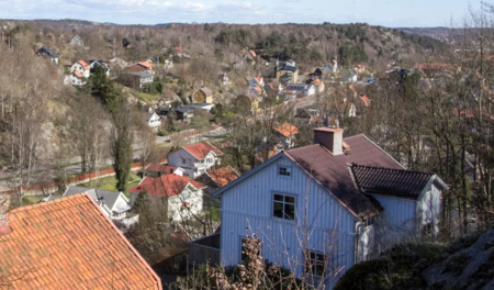Vy över ett bostadsområde med många hus, träd och kuperad terräng under en klar himmel.