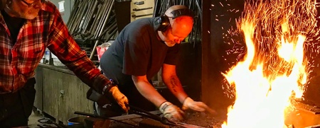 Två personer arbetar vid en smedja med eld och gnistor, bär hörselskydd och skyddsglasögon.