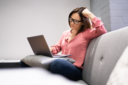 Kvinna med glasögon sitter på soffa och arbetar på en bärbar dator, ser fundersam ut.