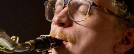 Närbild på en person med glasögon och mustasch som spelar saxofon.