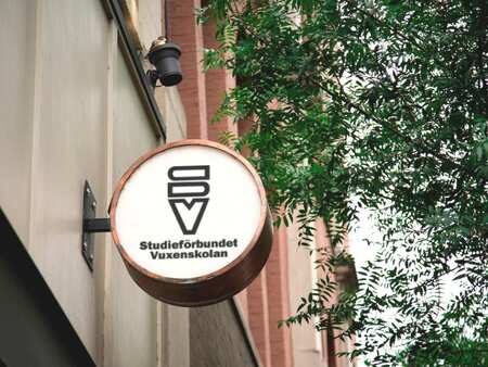 En rund ljusskylt med Studieförbundet Vuxenskolans logotyp på en husvägg med träd i bakgrunden.