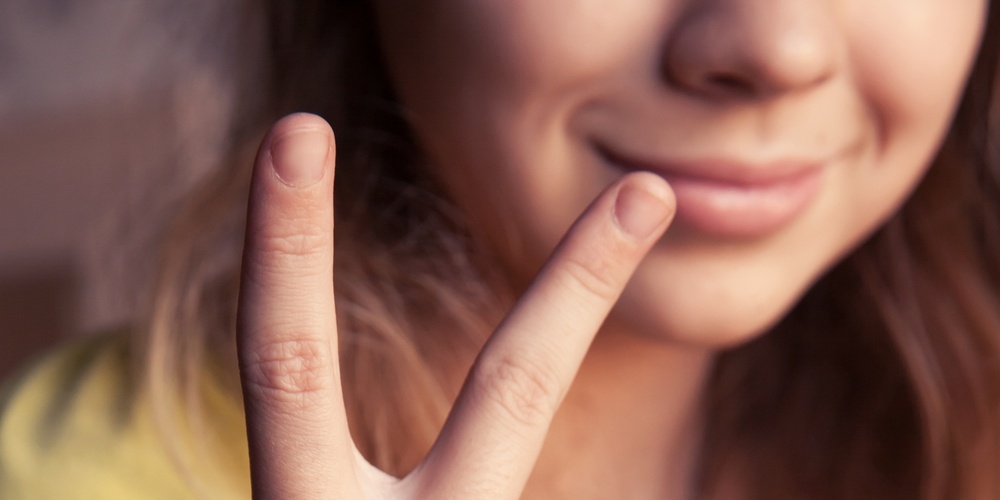En person i gul tröja visar ett V-tecken med handen och har färgglada armband på handleden.