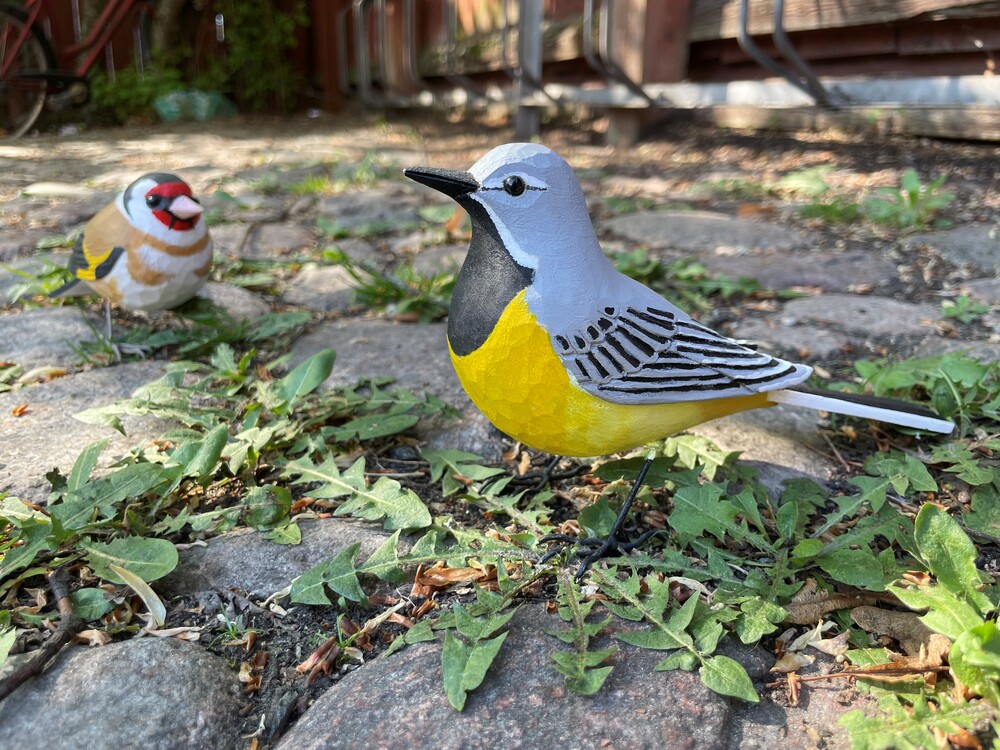 Två färgglada fågelfigurer av trä står på en stenig mark med gröna växter.