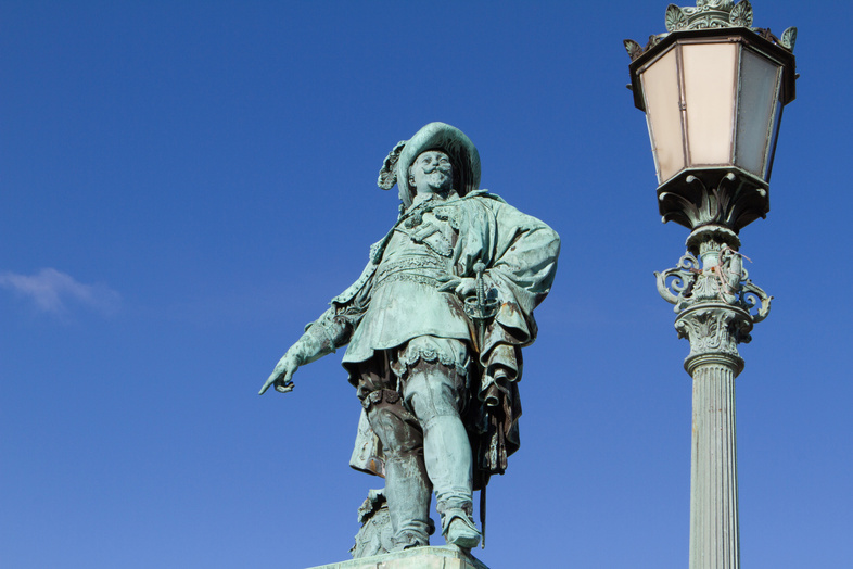 Staty av en man i historiska kläder bredvid en gammaldags gatlykta mot blå himmel.