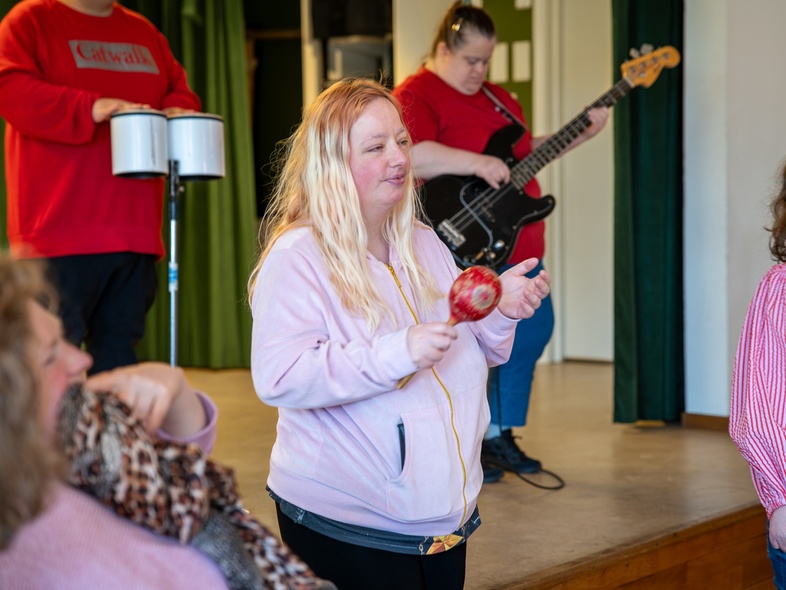 En grupp personer spelar musik tillsammans på en scen, med instrument som maracas, bas och bongotrummor.