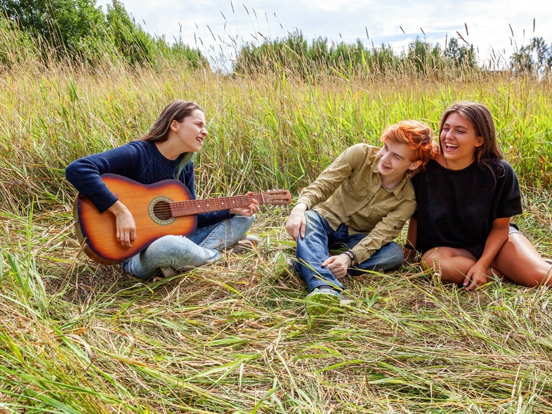 Tre unga personer sitter i gräset, en spelar gitarr och de andra två lyssnar och skrattar.