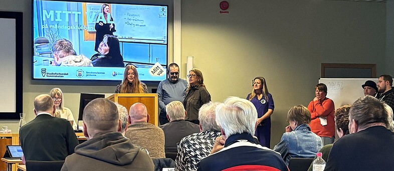 Möte eller presentation i konferensrum med flera personer, en talare vid podiet och publik som lyssnar.