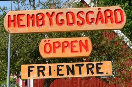 Träskyltar med texten 'Hembygdsgård', 'Öppen' och 'Fri entré' framför ett rött hus och gröna träd.