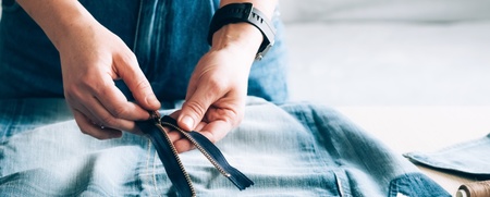 Person arbetar med att sy om ett par jeans, med dragkedja, måttband och sytråd på bordet.