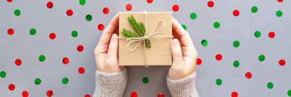 Två händer håller ett inslaget paket med snöre och grankvist mot en bakgrund med röda och gröna prickar.