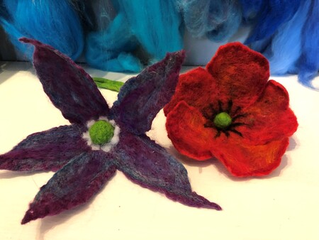 Två tovade blommor, en lila med fem kronblad och en röd med svart mitt, ligger på ett bord.