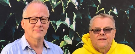 Två äldre män står bredvid varandra utomhus framför en vägg täckt av gröna blad.