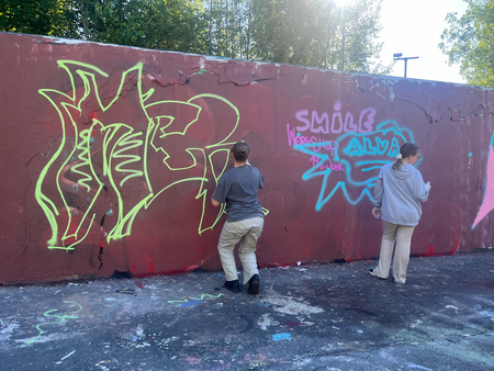 Två personer målar graffiti på en röd mur utomhus, omgivna av träd och dagsljus.
