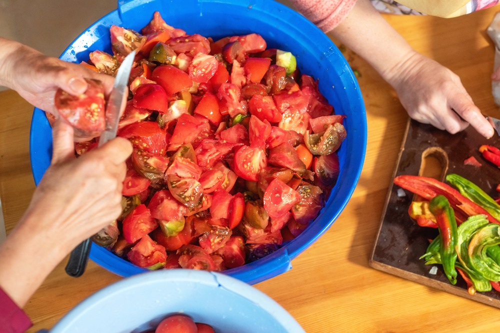 Två personer skär tomater och paprika vid ett bord, med en stor blå skål fylld med hackade tomater.