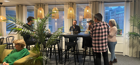 Flera personer samtalar och fikar i ett ljust kontorslounge med stora fönster, gröna växter och hängande lampor.