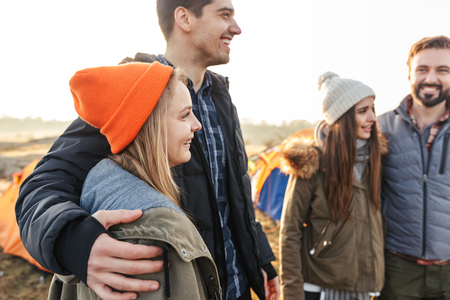Fyra unga vuxna står tillsammans utomhus vid tält, klädda i varma kläder och mössor, och ser glada ut.