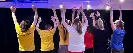 Sju personer står med ryggen mot kameran och armarna uppsträckta i ett upplyst rum med trägolv.