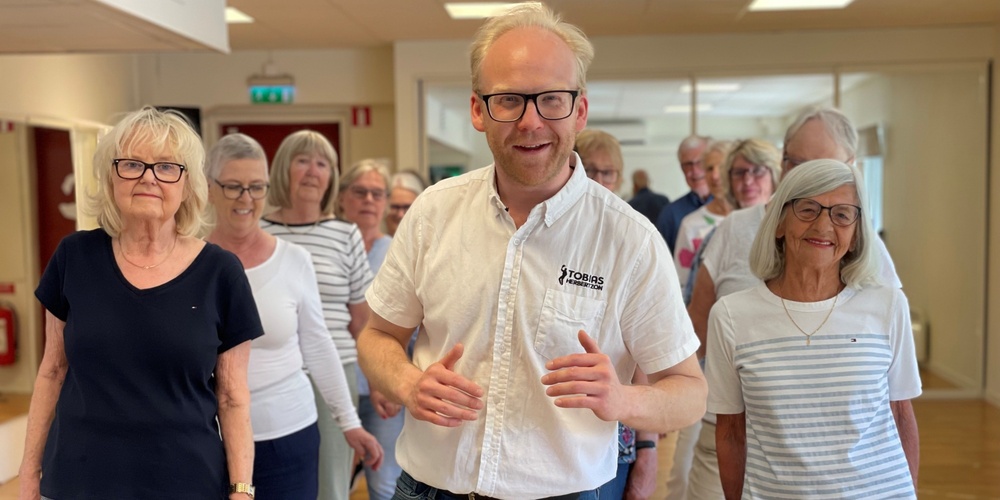 En grupp äldre personer och en yngre man står tillsammans inomhus, leende och avslappnade.