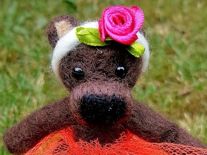 En liten brun nalle i röd tyllkjol och blomdiadem sitter på en vit sten på gräsmatta.