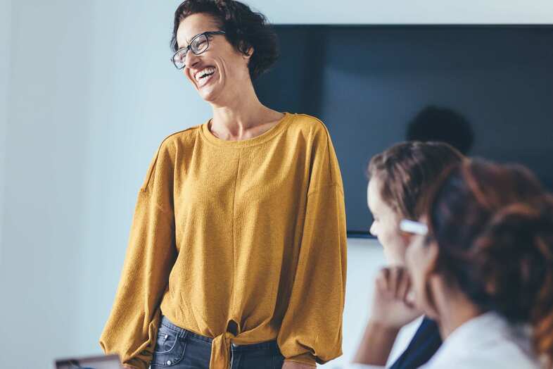 En leende person med gul tröja står framför en stor svart skärm medan två personer sitter och tittar på personen.