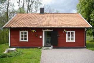 Rött trähus med vita knutar, två svenska flaggor och grusgång framför, omgiven av grönska.