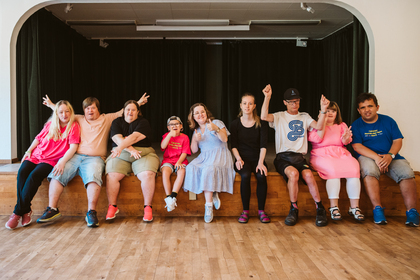 En grupp unga personer sitter på en scenkant och poserar glatt tillsammans inomhus.