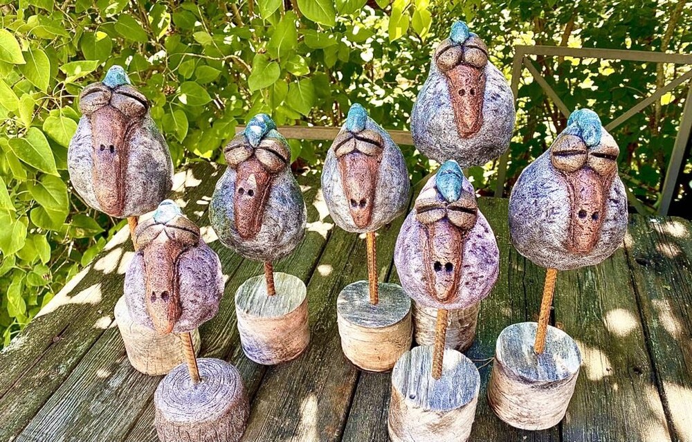 Flera färgglada fågelliknande skulpturer av betong står på ett träbord utomhus bland gröna blad.